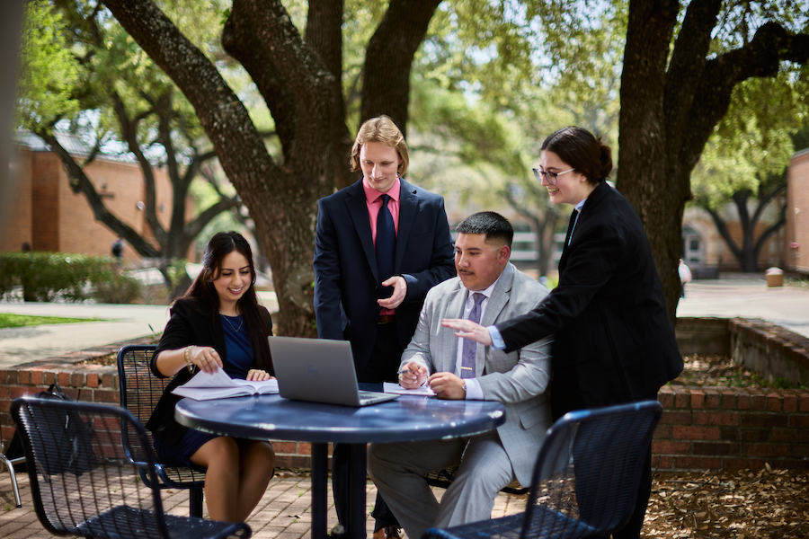 four law students stand and sit together around a table outside and look at a laptop screen