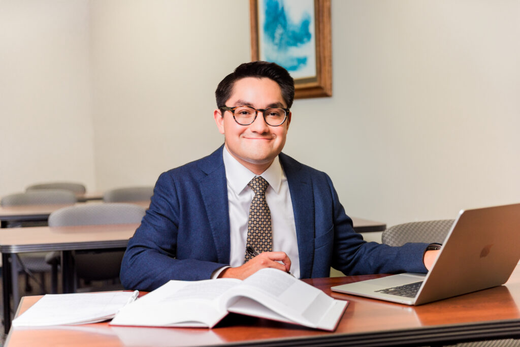 Student wearing a blue suit smiles. An open book and laptop is on the desk in front of him