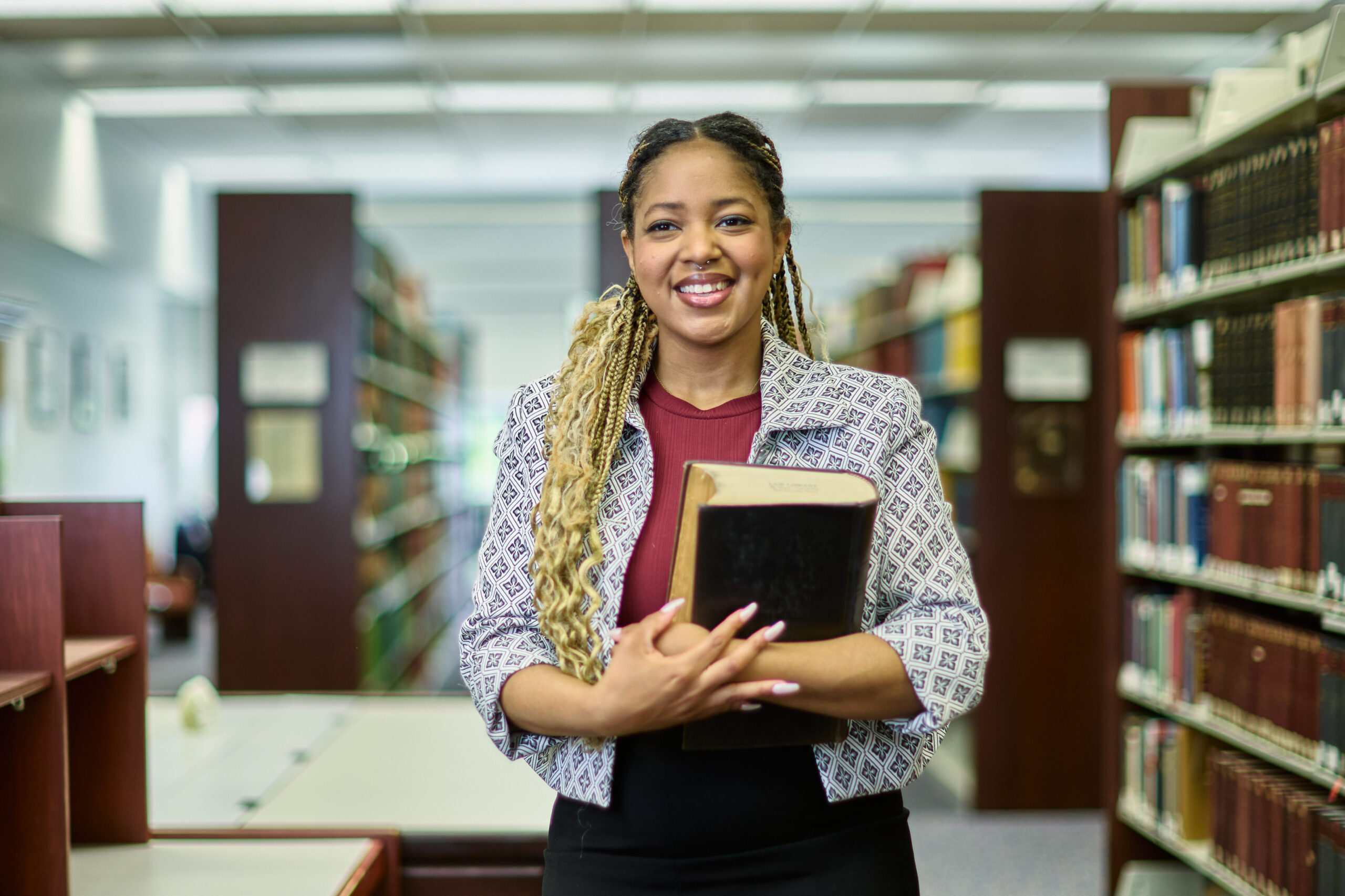 Female Law student holding a pile of books smiles as she stans in by a bookshelf in the library.