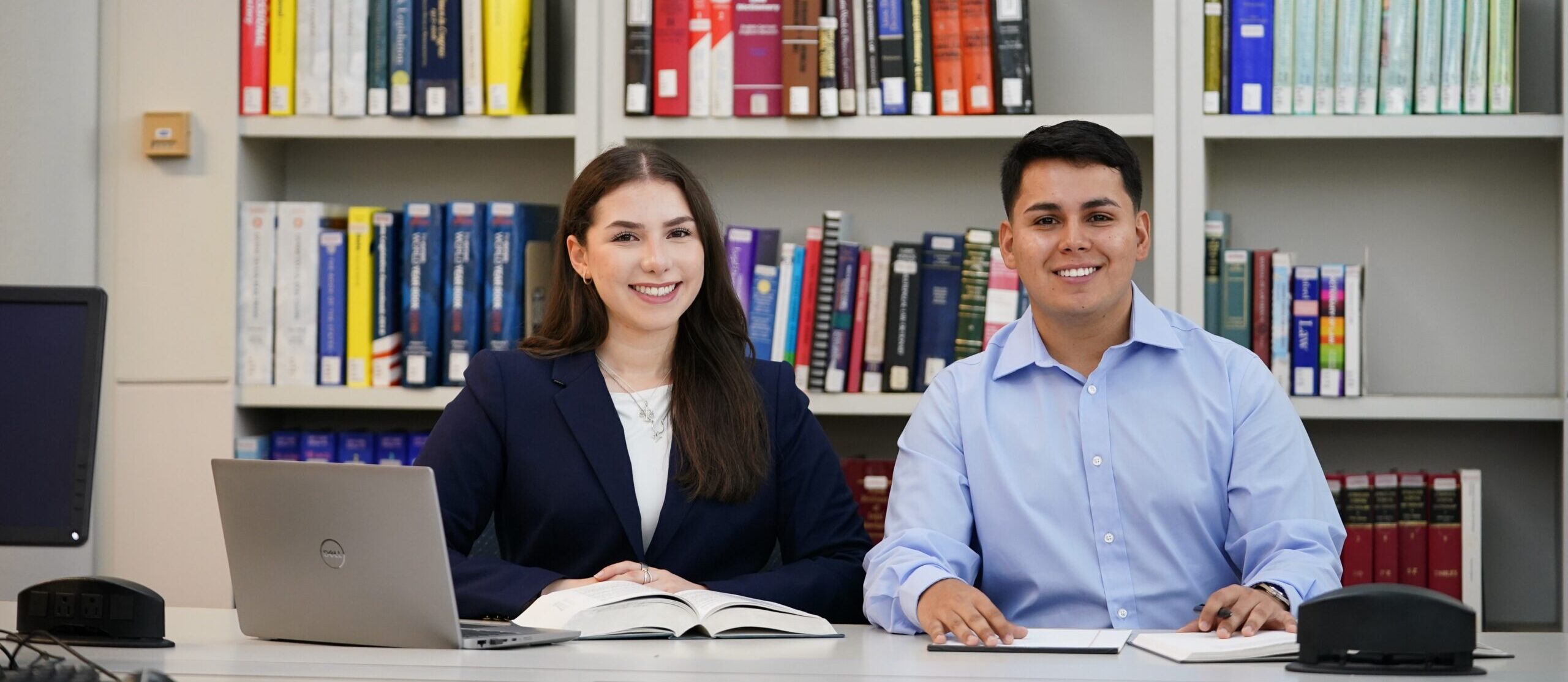Two students smile as they study together in the library. On the table in front of them a laptop and two books can be seen. Behind them is a large bookshelf filled with many books.