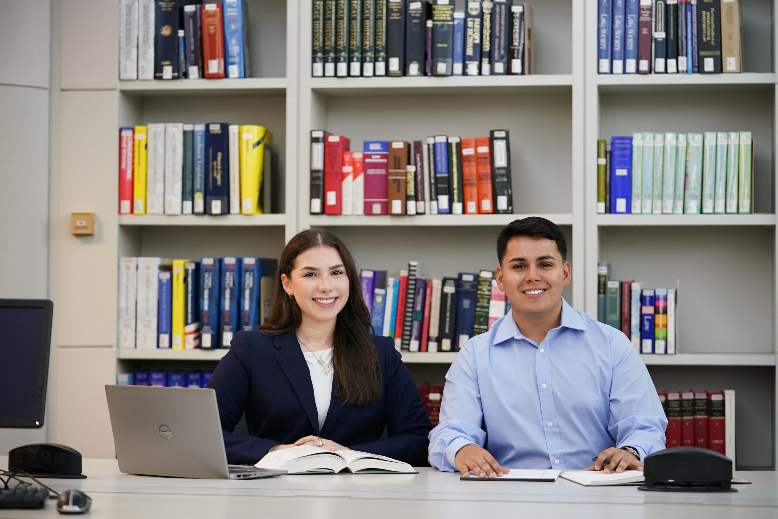 Two students smile as they study together in the library. On the table in front of them a laptop and two books can be seen. Behind them is a large bookshelf filled with many books.