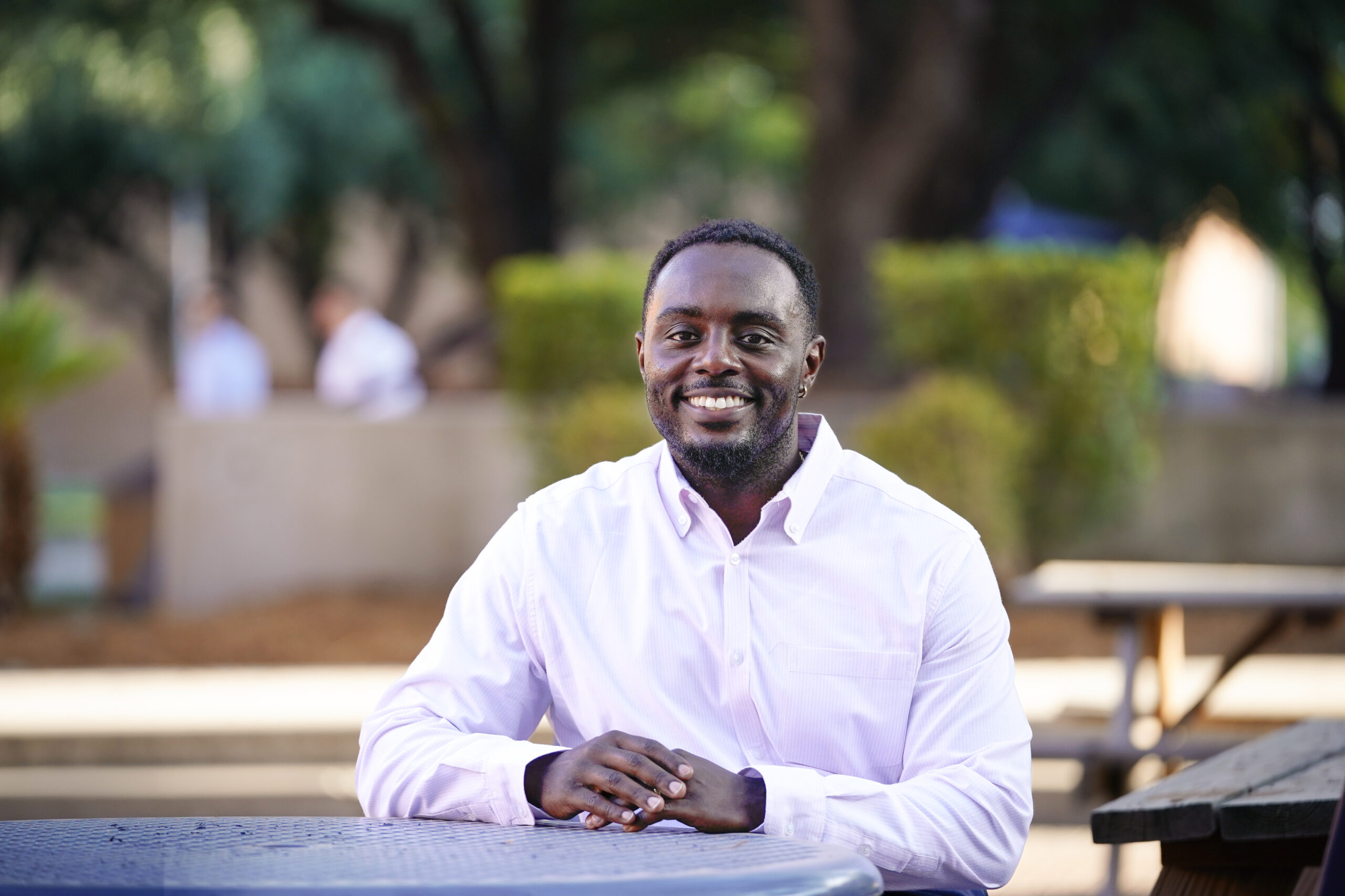 Smiling male student wearing a white shirt sits at a table outside.