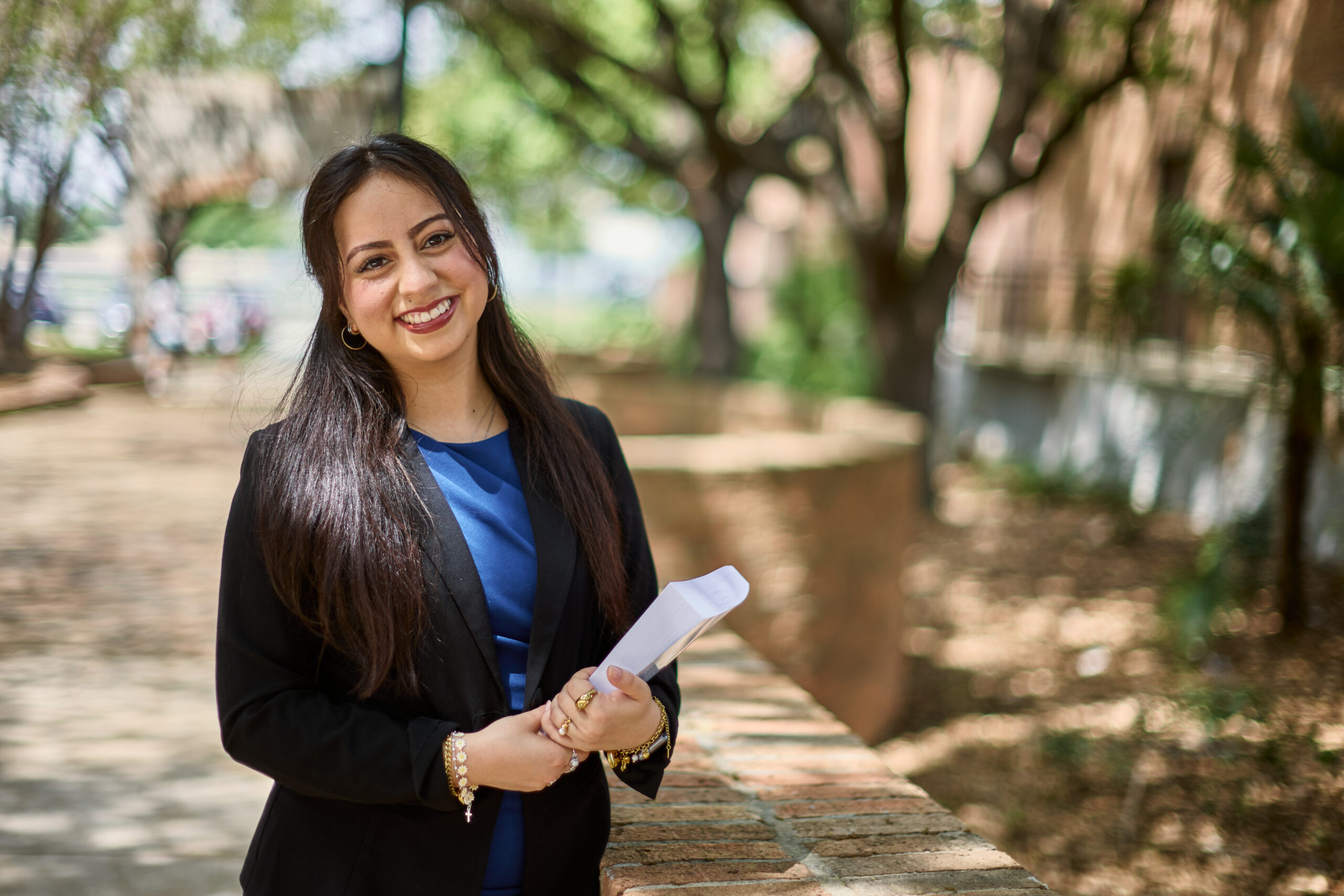 Smiling female student wearing a suit jacker and blue dress holding a book.